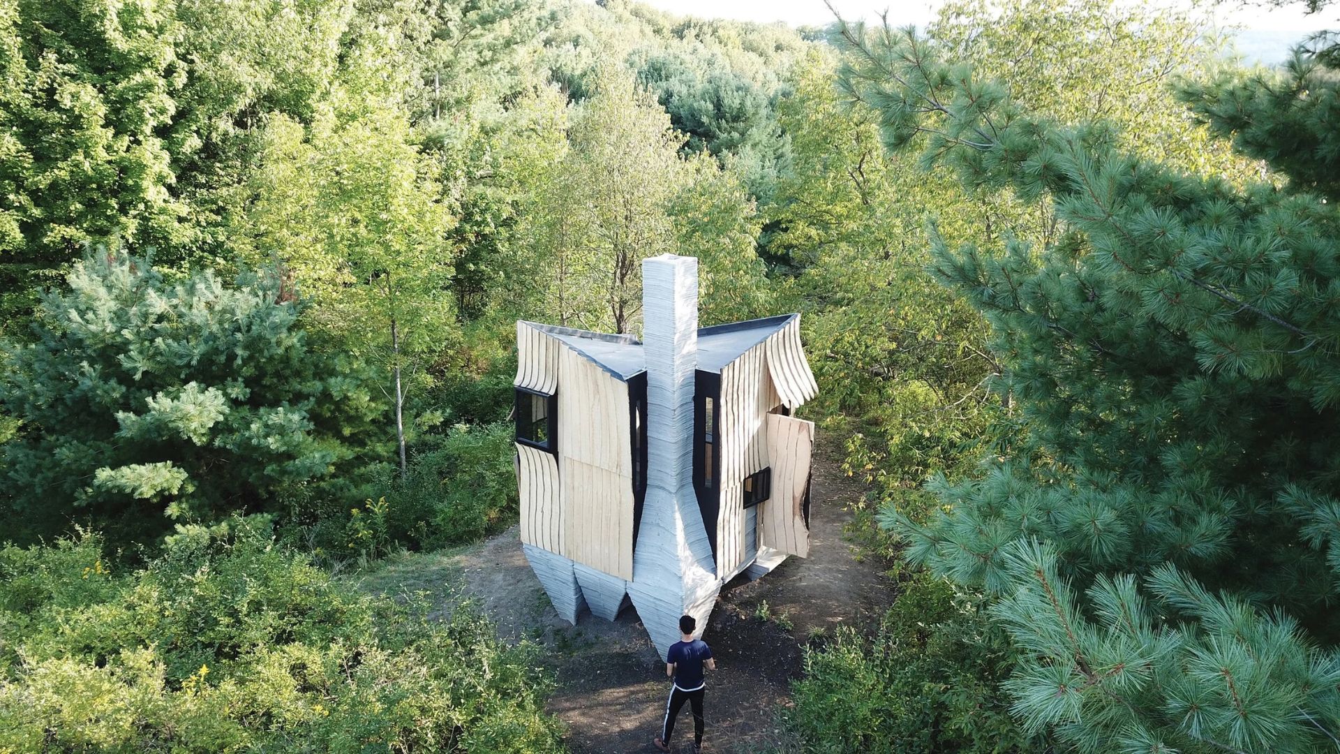 A larger than life, sculptural architectural installation stands in a wooded landscape, with layered light-colored surfaces, dark openings, and a central vertical element. A person stands in front of the structure for scale.