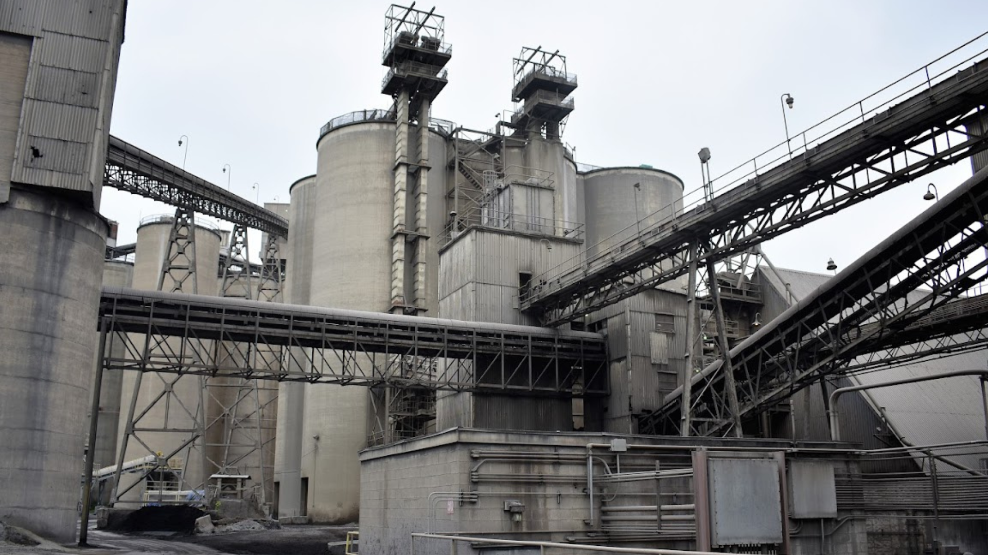 Industrial facility with large cylindrical silos, metal structures, and elevated conveyor systems interconnected across the site. The gray concrete and steel buildings appear weathered, with pipes, walkways, and machinery visible throughout the complex under an overcast sky.