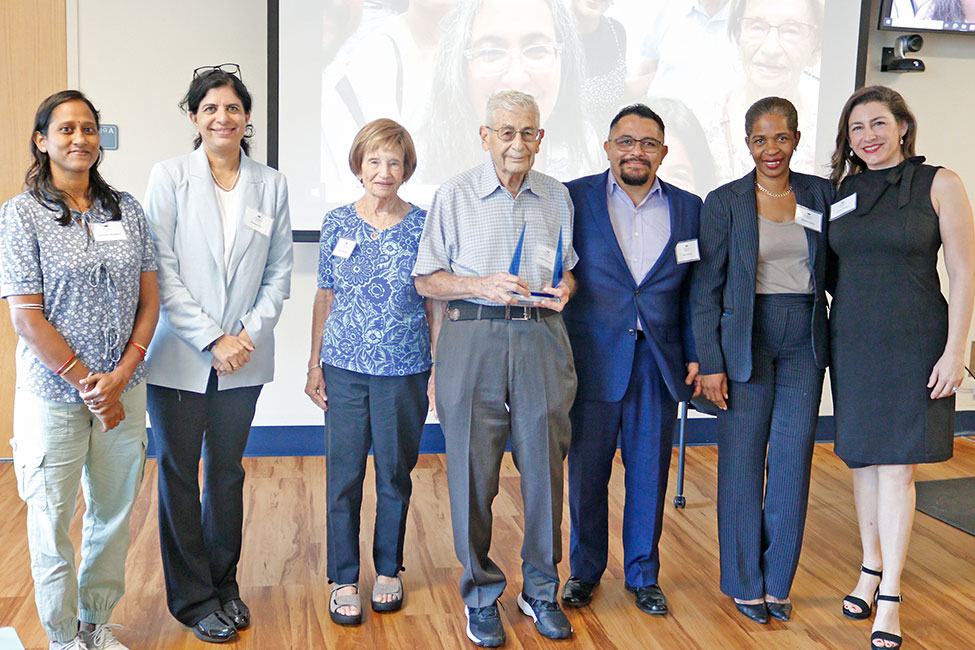 Group of people stands in a line smiling at the camera in an indoor classroom setting with a projector screen in the background. The person in the center holds a clear, glass award.