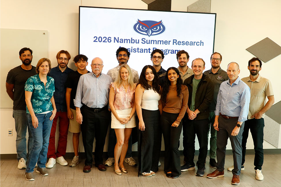 Group photo of about 15 people standing in two rows in an indoor room with wood-look flooring. Behind them is a large screen displaying an owl logo and the text
