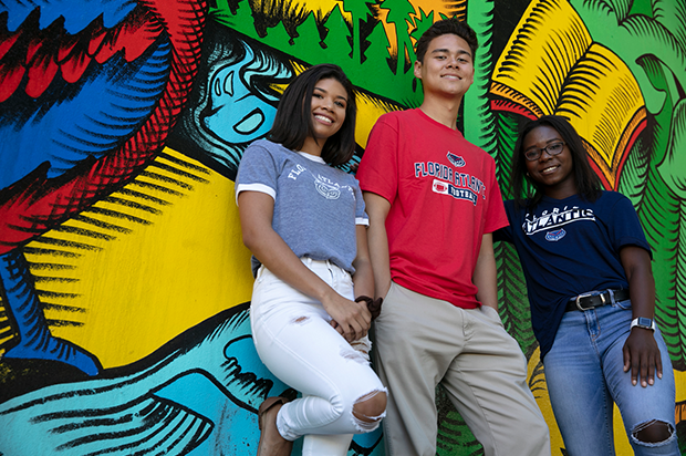 A male student and two female student lean against a wall covered with art