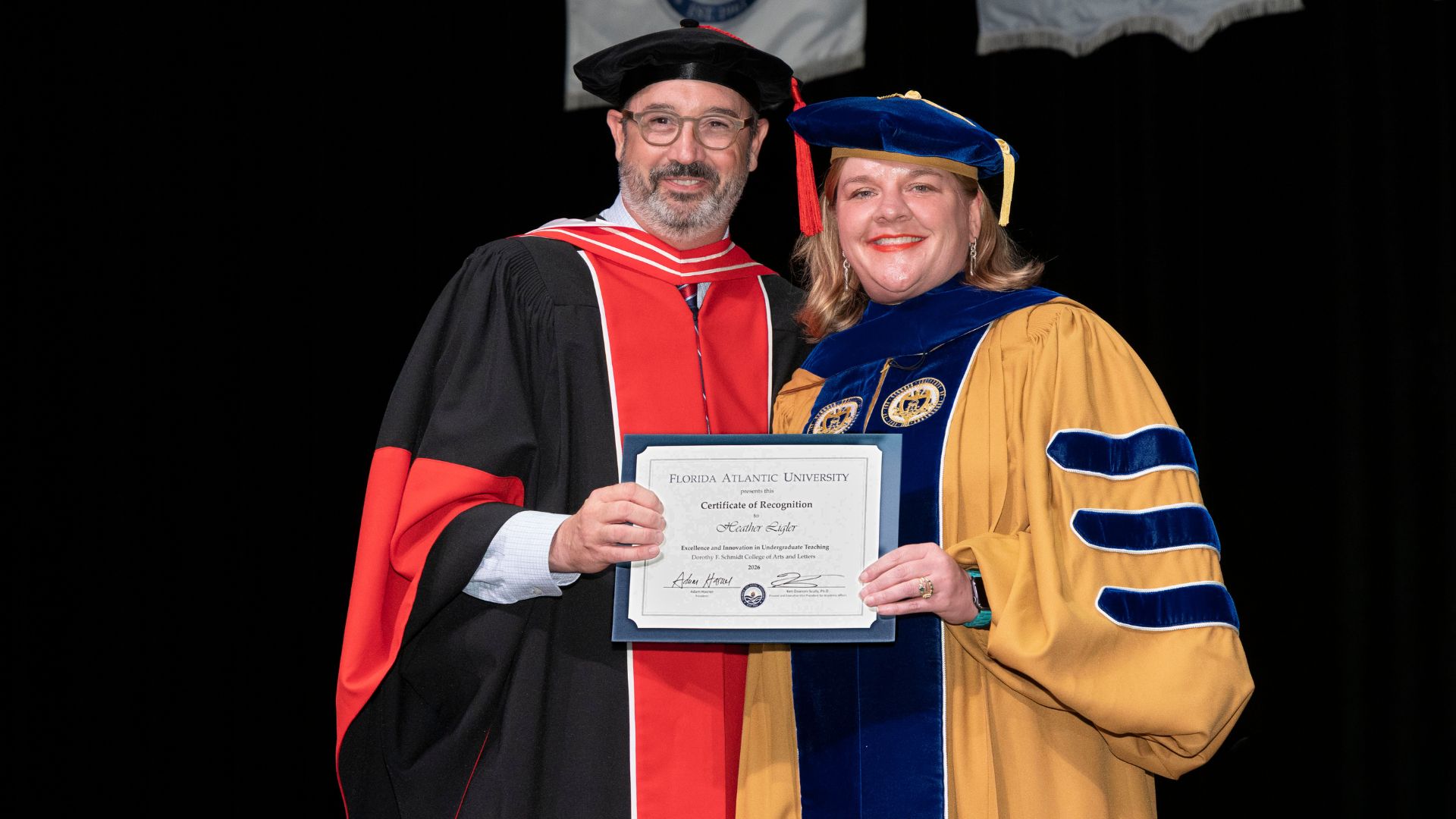 Two people in academic regalia pose while holding a Florida Atlantic University certificate of recognition for Excellence and Innovation in Undergraduate Teaching.