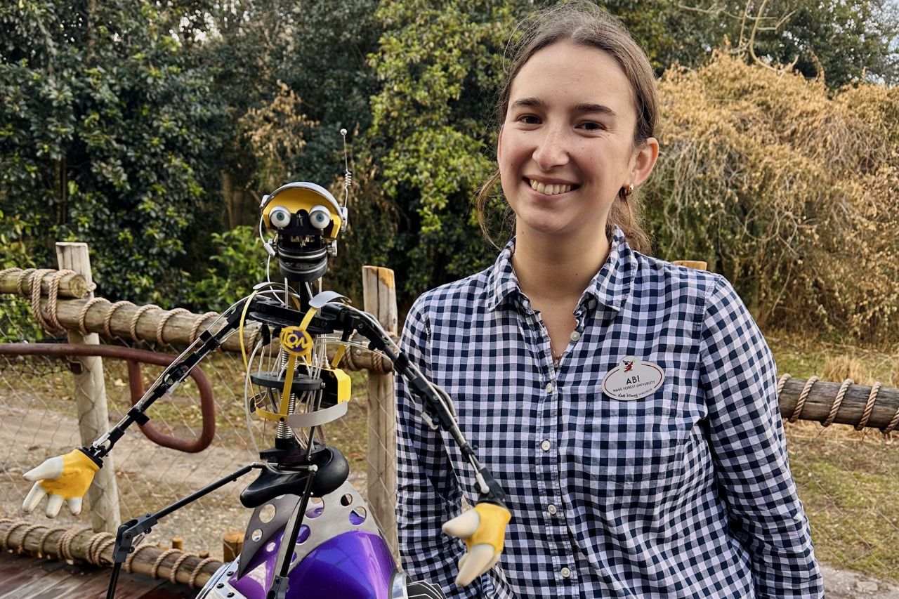 A person stands next to a robot with yellow accents on its eyes and hands. The person is wearing a black and white checkered shirt and a nametag. They are both smiling at the camera in an outdoor setting with trees in the background.