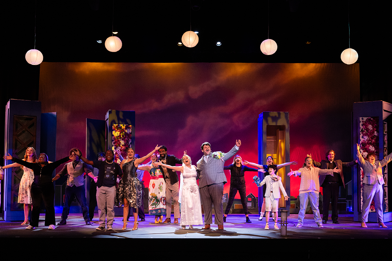 Cast shot of actors singing with there arms raised. there is colorful abstract decoration behind them on the stage