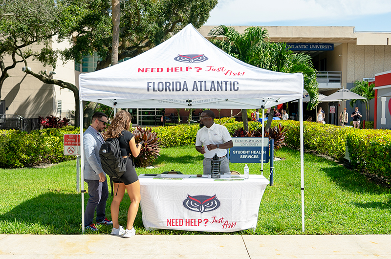 a white tent with FAU logos on it placed on the grass next to a busy walkway on campus, with staff members helping students find their way