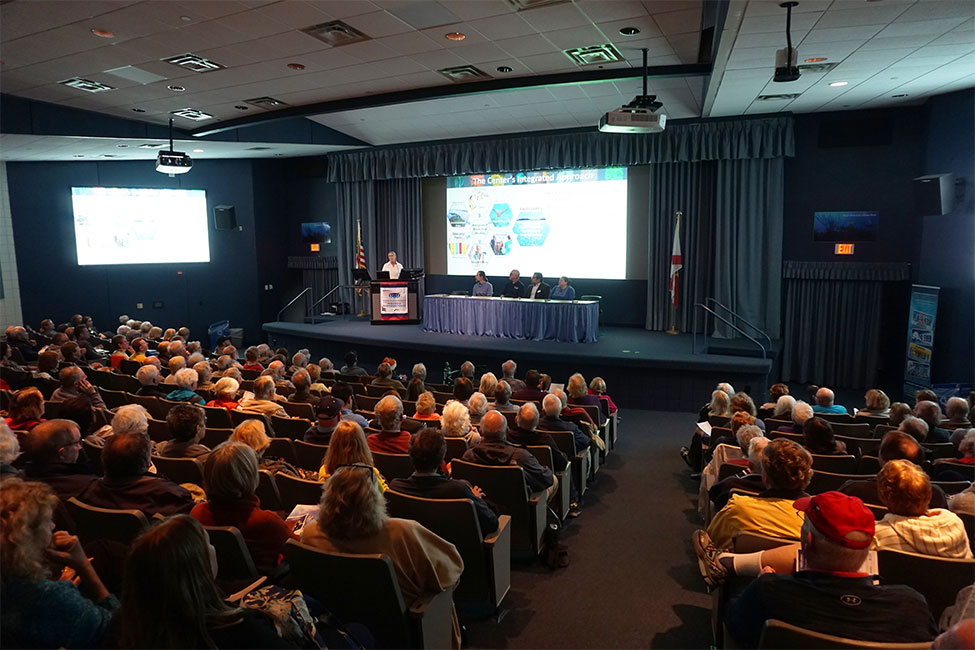 Photo of large, tiered lecture hall. Seats are filled with attendees. A person stands behind a podium. A table is set in the middle of the stage with four panelists behind. A large screen with a colorful Powerpoint slide is against the wall behind the panelists.