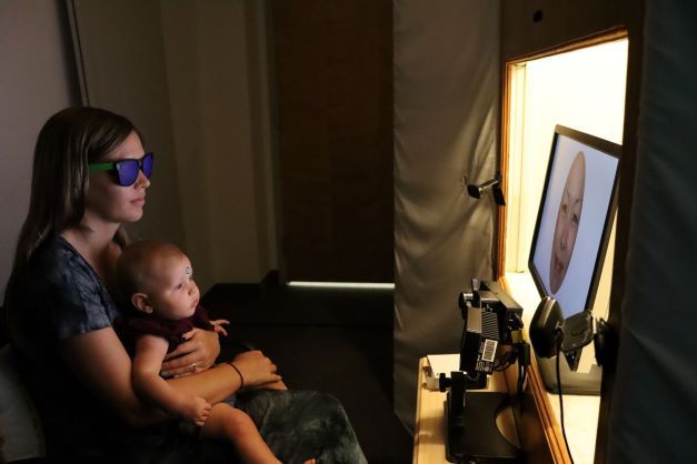 Infant seated on mother's lap in front of a video screen. A small camera track's the baby's eye gaze.