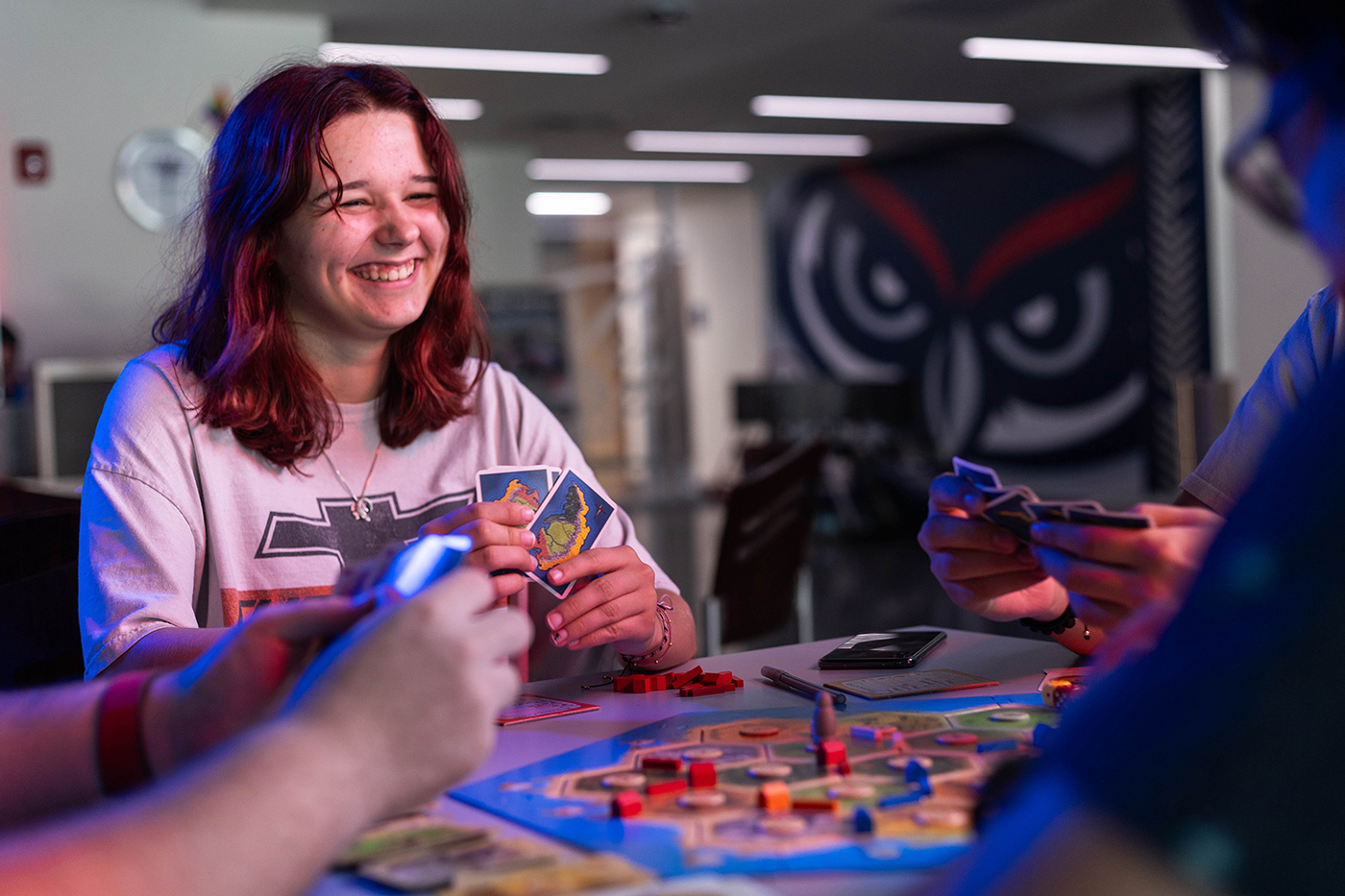 a young lady smiling with cards in hand as she plays a colorful, stragetic board game with others